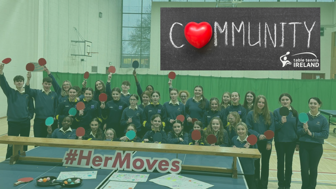 A large group of schoolgirls in uniform proudly hold up table tennis paddles behind a table displaying posters and a sign that reads "#HerMoves" in a school gym.