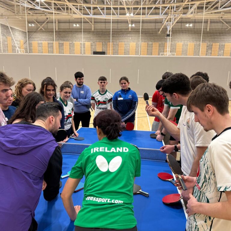 A group of young adults gather around a table tennis table, listening attentively as a coach in a green 'IRELAND' shirt demonstrates a technique.