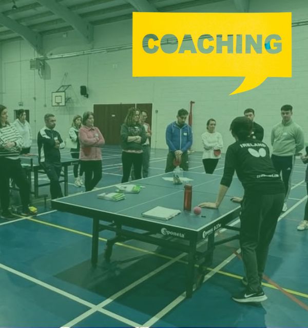 A group of adults gathered around a table tennis table during a coaching session in a sports hall.