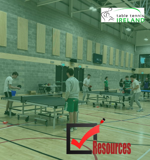 Players of various ages compete in table tennis matches inside a large indoor sports hall with multiple tables and a viewing gallery above.