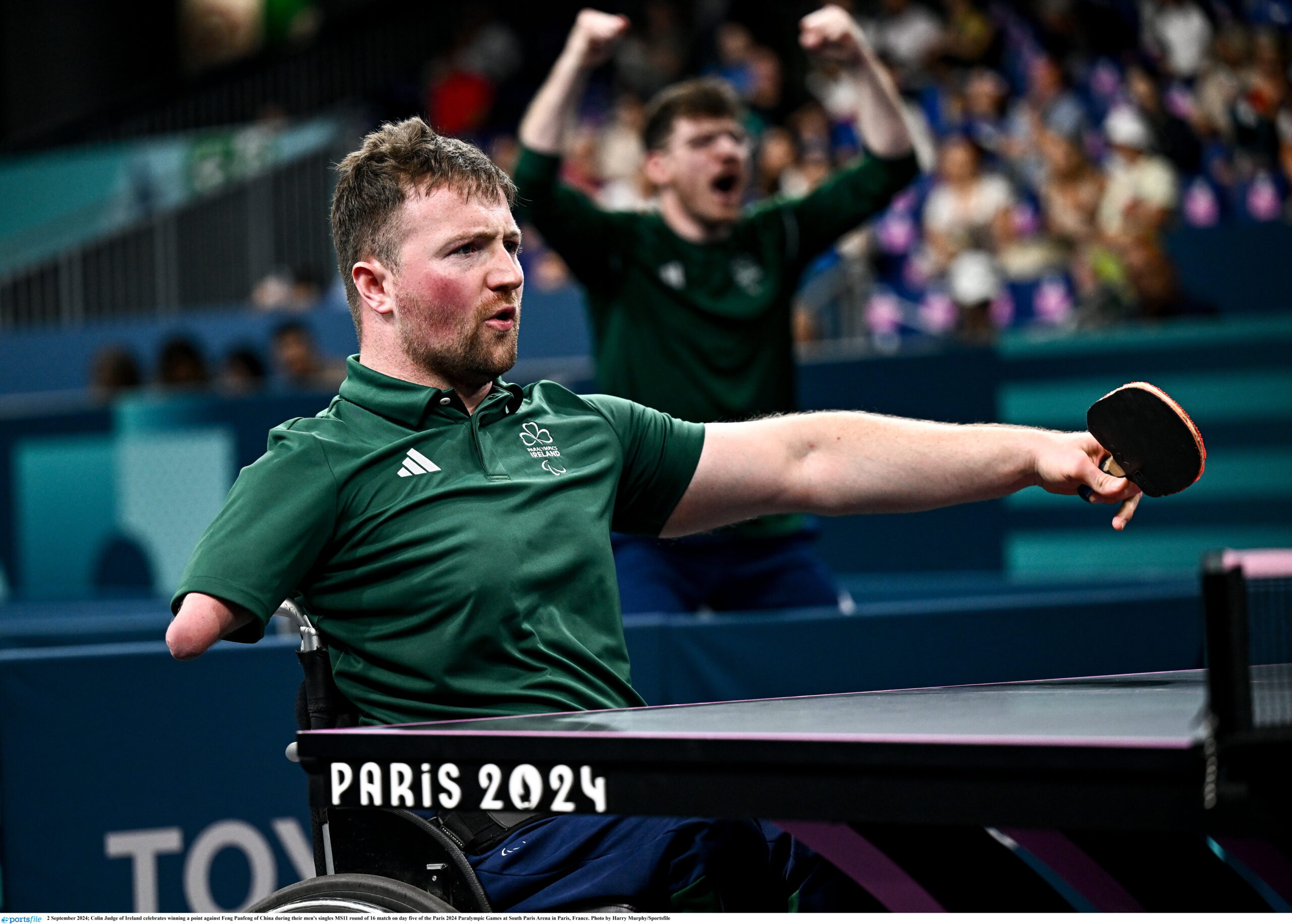 Irish Paralympic table tennis player Colin Judge competes at Paris 2024, striking the ball with an outstretched arm while seated in his wheelchair. He wears a green Ireland team shirt, and a teammate celebrates in the background as the crowd watches from the stands.