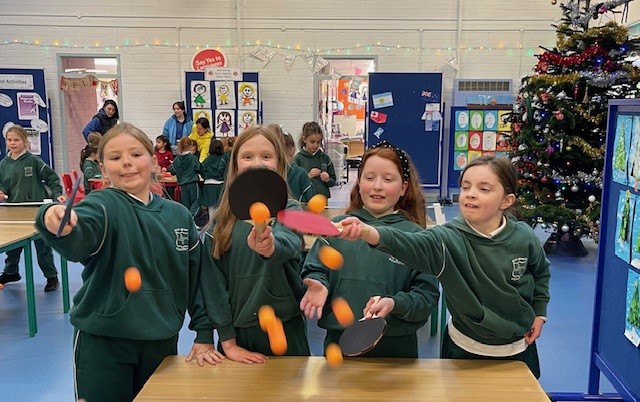 Four primary school girls in green uniforms play table tennis indoors at a national school in Cork, tossing orange ping pong balls into the air while holding paddles. Other children and classroom displays are visible in the background, along with a decorated Christmas tree.