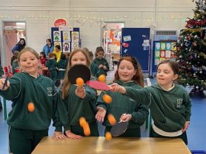 Four primary school girls in green uniforms play table tennis indoors at a national school in Cork, tossing orange ping pong balls into the air while holding paddles. Other children and classroom displays are visible in the background, along with a decorated Christmas tree.