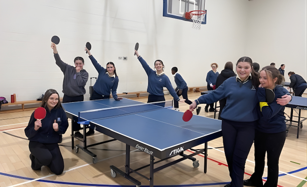 Schoolgirls smiling and posing around a table tennis table in a gym, holding paddles and enjoying the session.